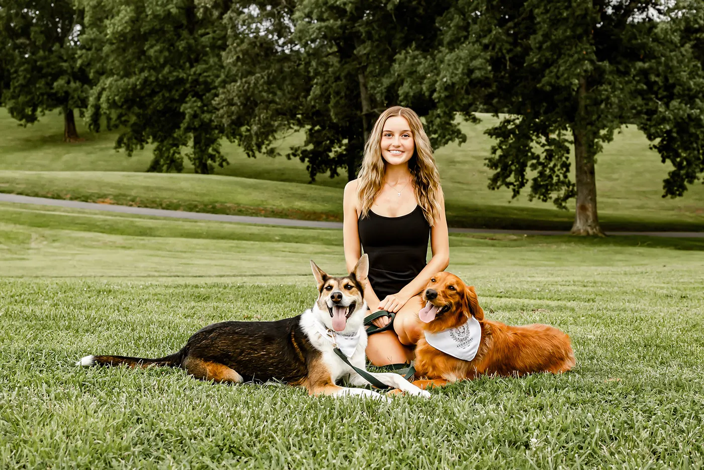 Avery sitting on the grass with a shepherd mix and a golden retriever, both wearing Averyday bandanas